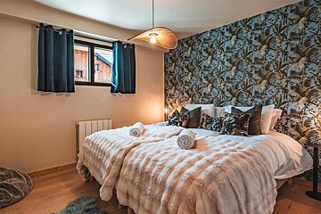 A cozy bedroom with a fluffy bed, tropical leaf accent wall, unique woven pendant light, and a window with blue curtains overlooking wooden buildings.