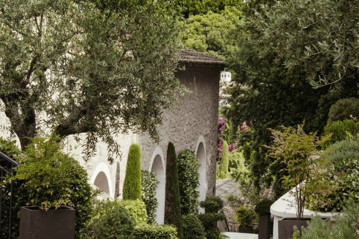 A stone building with arches is surrounded by trees and bushes