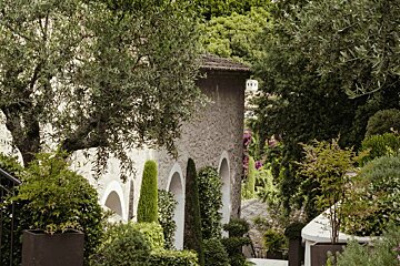 A stone building with arches is surrounded by trees and bushes