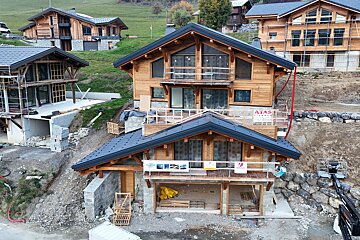 Multiple modern wooden chalets are under construction on a green, sloped hillside. Some appear complete, while others show exposed foundations and scaffolding.