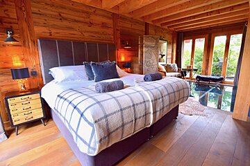 A cozy, wood-paneled bedroom with a large bed and plaid blanket, featuring a rustic yet modern design and large windows overlooking lush greenery.