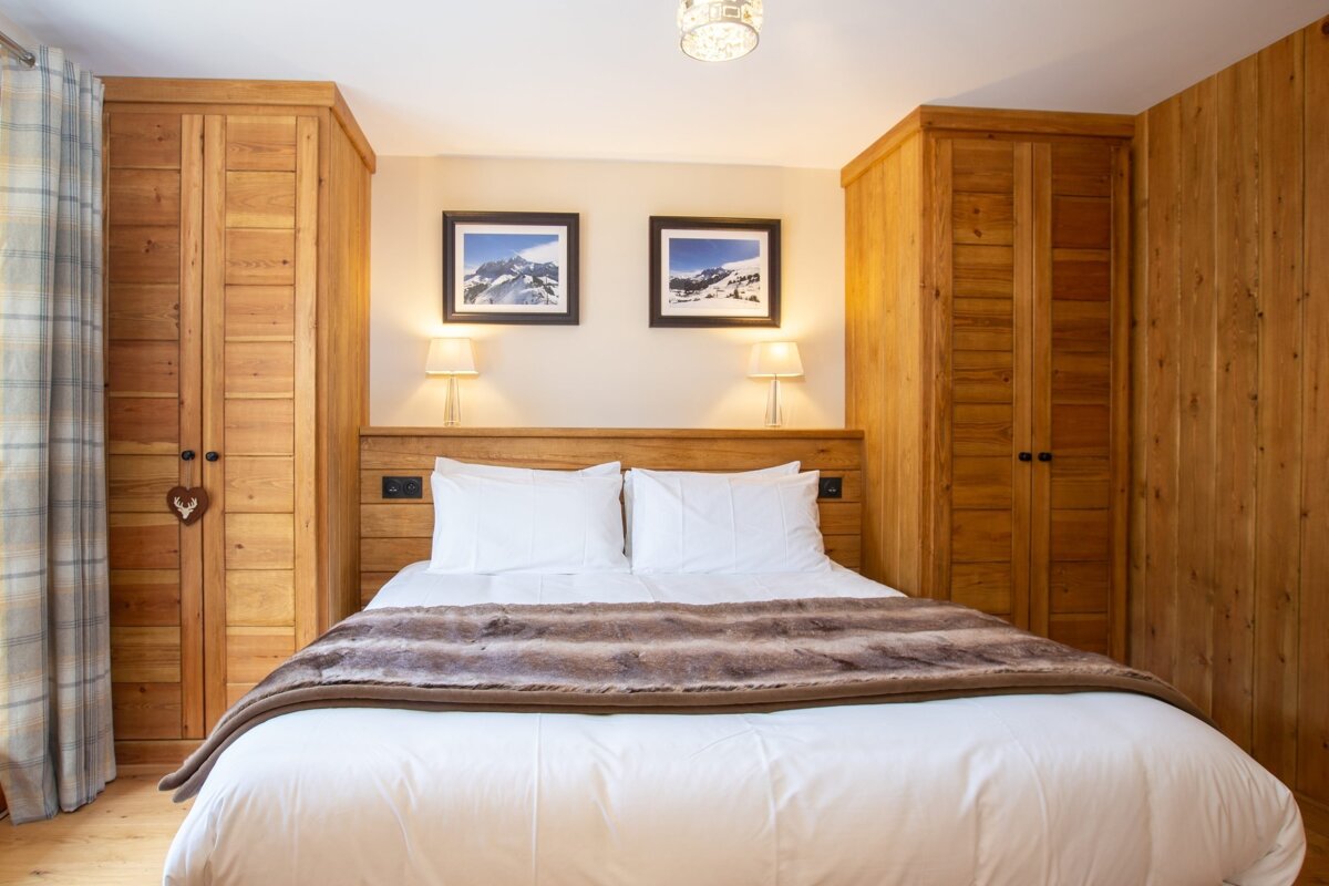 A cozy, wood-paneled bedroom featuring a large bed with white linens and a faux fur throw, flanked by built-in wooden wardrobes. Mountain art hangs above the bed, illuminated by warm lamps.