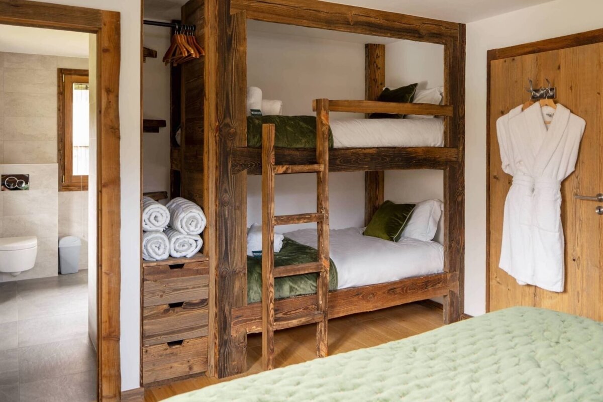 Rustic wooden bunk beds with green accents, rolled towels, and a white robe create a cozy, functional lodging space, adjacent to a modern bathroom.