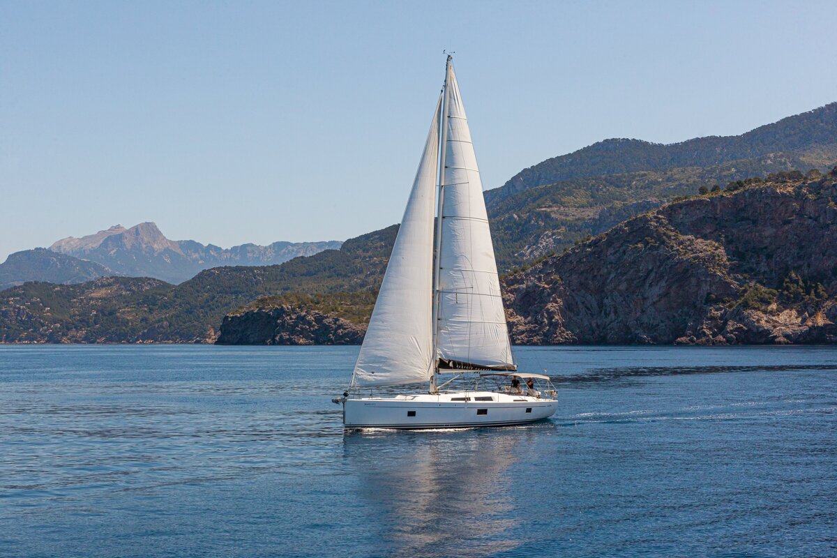 A white sailboat glides across calm blue water, framed by rugged mountains under a clear sky.
