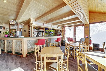 Cozy ski lodge bar/restaurant with rustic wooden decor, stone bar, dining tables, and large windows overlooking a snowy mountain view.