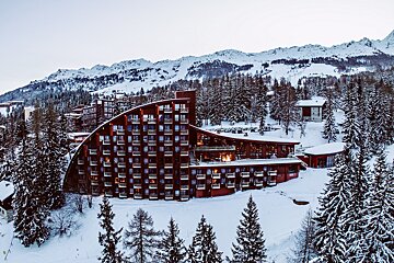 A large, curved ski resort building stands amidst a snowy mountain landscape, surrounded by snow-covered pine trees under a pale sky.