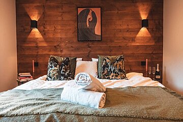 A cozy bedroom featuring a wooden wall, soft bed with tropical pillows, ambient lighting, rolled towels, and a romantic setup with champagne.