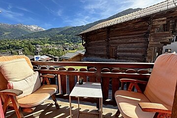 A balcony with two wooden chairs and a table overlooking a Swiss mountain village and log cabins under a blue sky.