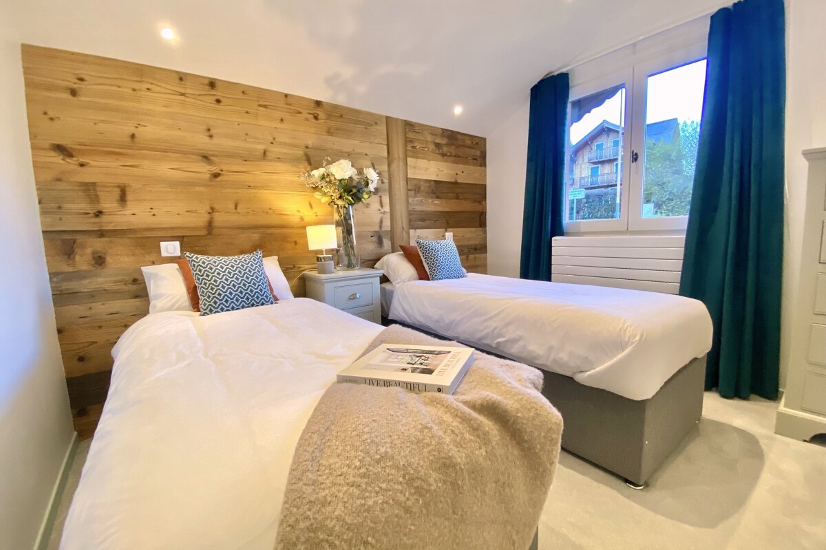 A cozy twin bedroom featuring a rustic wooden accent wall, two beds with white linens, a shared nightstand, and blue curtains by the window.