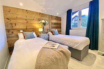 A cozy twin bedroom featuring a rustic wooden accent wall, two beds with white linens, a shared nightstand, and blue curtains by the window.