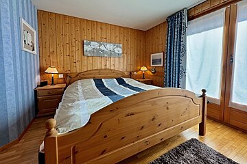 Cozy bedroom with wood panel walls, a large wooden bed with a blue/white striped duvet, nightstands, lamps, and a window with patterned blue curtains.