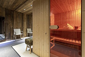 A rustic wood-paneled spa area features a glowing red sauna with benches and towels, next to a seating area with modern chairs and a furry stool.