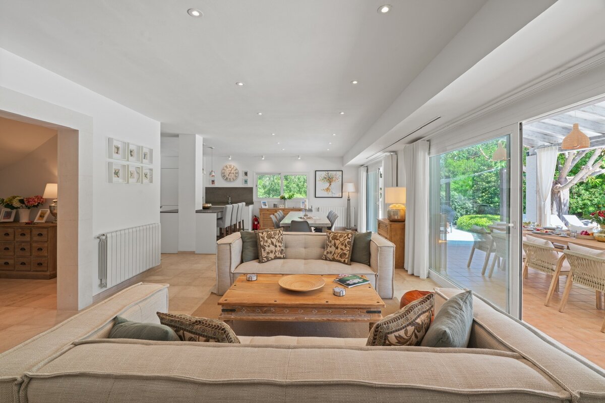 Bright, open-plan living room with sofas, a wooden coffee table, connecting to a kitchen, dining area, and an outdoor patio with seating.