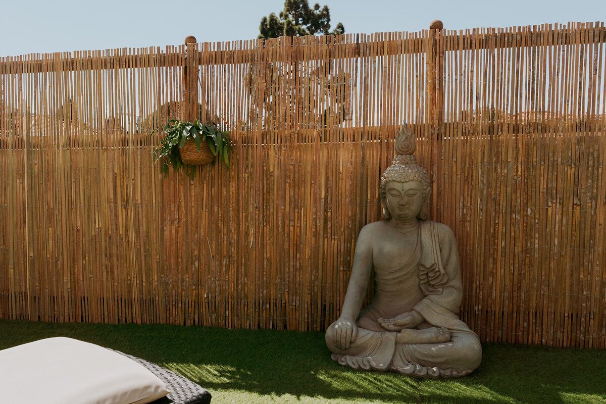 A stone Buddha statue rests on green grass next to a tall bamboo fence with a hanging plant. A lounge cushion is in the foreground.