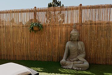 A stone Buddha statue rests on green grass next to a tall bamboo fence with a hanging plant. A lounge cushion is in the foreground.