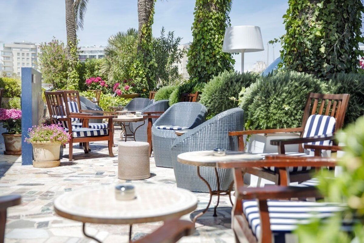 A charming outdoor patio with seating areas, including wooden and wicker chairs with blue-and-white striped cushions, surrounded by abundant foliage and blooming flowers.