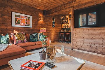 A cozy, wood-paneled living room with an orange sectional, vibrant cushions, modern art, a built-in bar, and a large TV. Rustic yet eclectic.