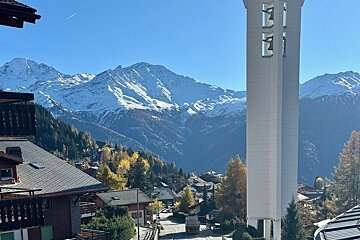 A scenic view of a mountain village featuring chalet roofs, a tall white bell tower, and majestic snow-capped peaks in the background.