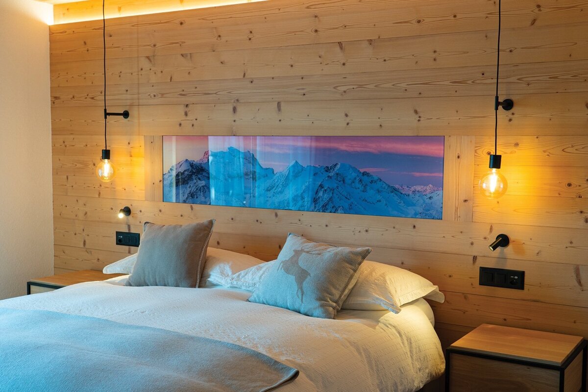 A cozy bedroom with a wooden accent wall, featuring a large backlit mountain view above the bed, illuminated by warm hanging lights.