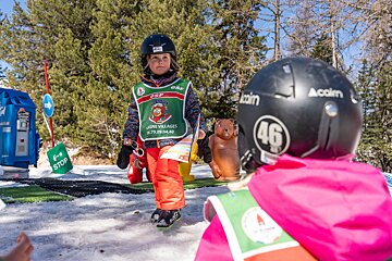Two young children in ski helmets and colorful gear are on a snowy mountain with trees. One faces the camera, holding sunglasses, while the other faces away.
