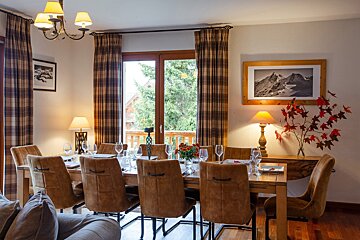 An inviting dining room features a long wooden table set for guests, brown chairs, plaid curtains, warm lighting, and a scenic window view.