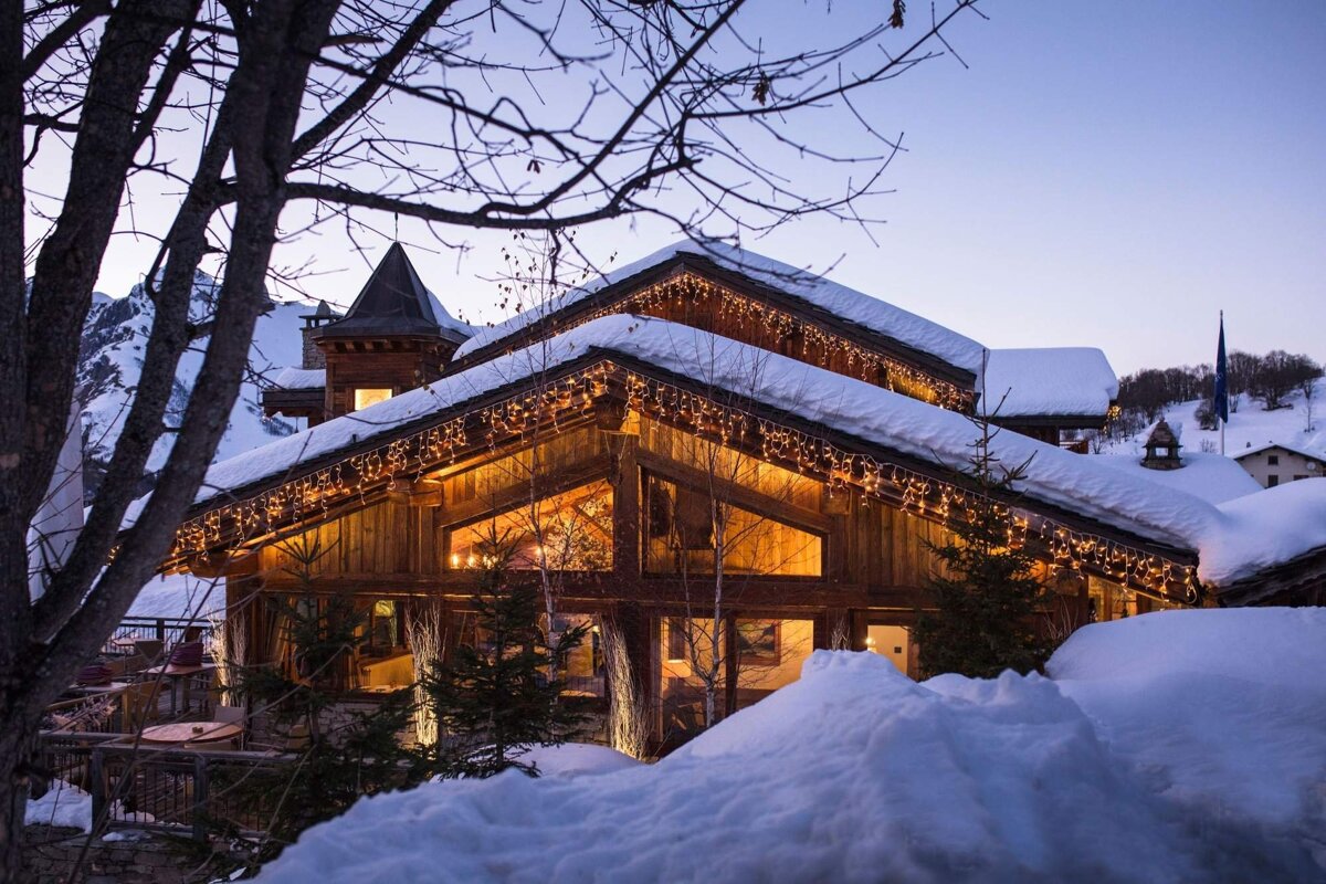 A cozy, snow-covered wooden chalet with warm lights glowing at dusk, set against a backdrop of mountains and deep winter snow.