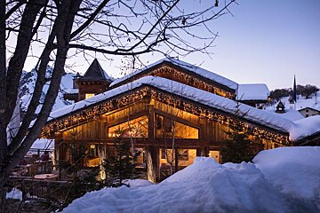 A cozy, snow-covered wooden chalet with warm lights glowing at dusk, set against a backdrop of mountains and deep winter snow.