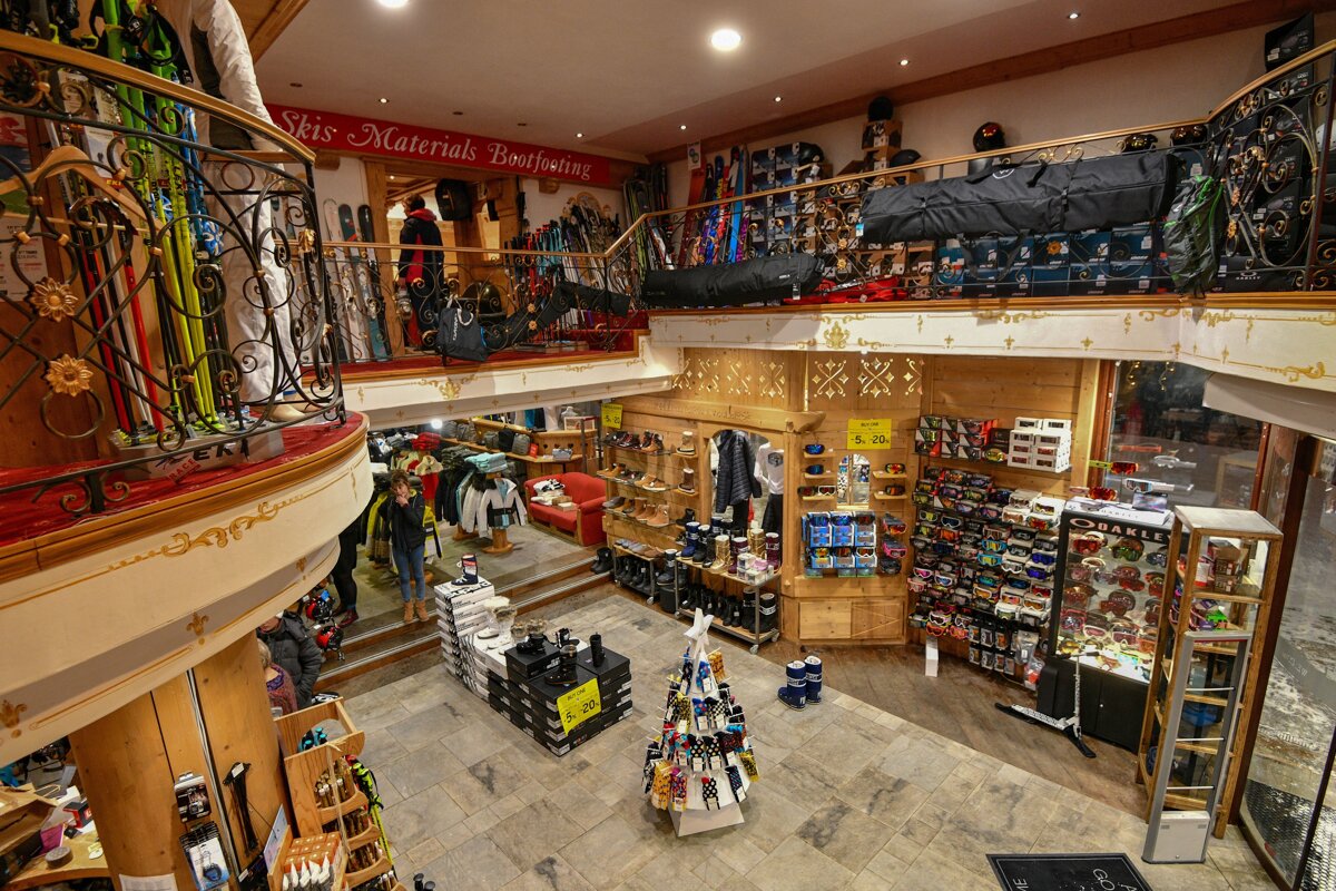 A cozy, two-story ski and bootfitting shop, richly decorated with wood and ornate railings, displays numerous skis, boots, goggles, and apparel, with customers browsing.