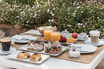 A vibrant outdoor breakfast spread with coffee, juice, pastries, open sandwiches, fruit, and yogurt, set among blooming white flowers.