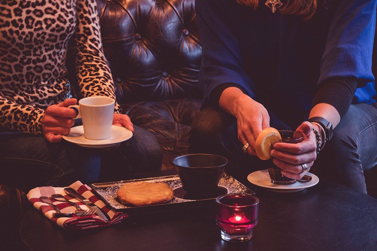 Two women sit on a couch drinking coffee and eating cookies