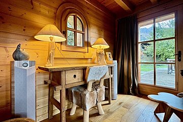 A cozy wooden room with a rustic desk, fur chair, arched window, and a door opening to an outdoor deck and green scenery.