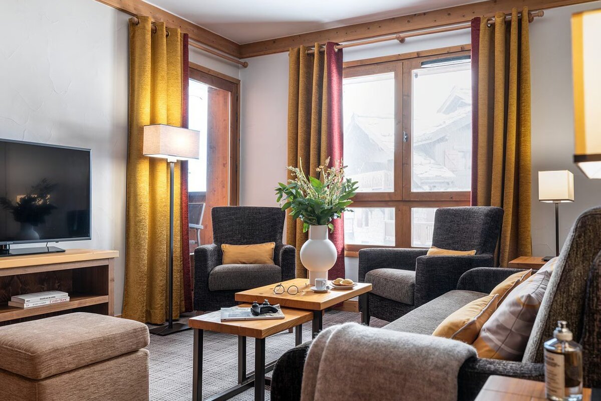 A cozy, modern cabin living room with grey armchairs, wooden tables, and yellow curtains framing a snowy window view.