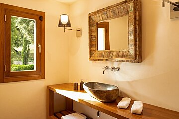 A rustic bathroom features a stone basin on a wooden counter, a reclaimed wood mirror, and a window overlooking a lush green garden.