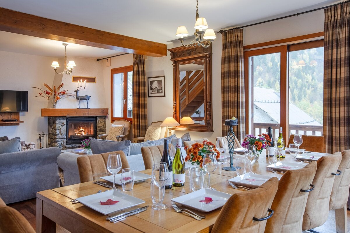 A cozy, rustic chalet interior featuring a long dining table, a warm fireplace in the living area, and large windows with a scenic mountain view.