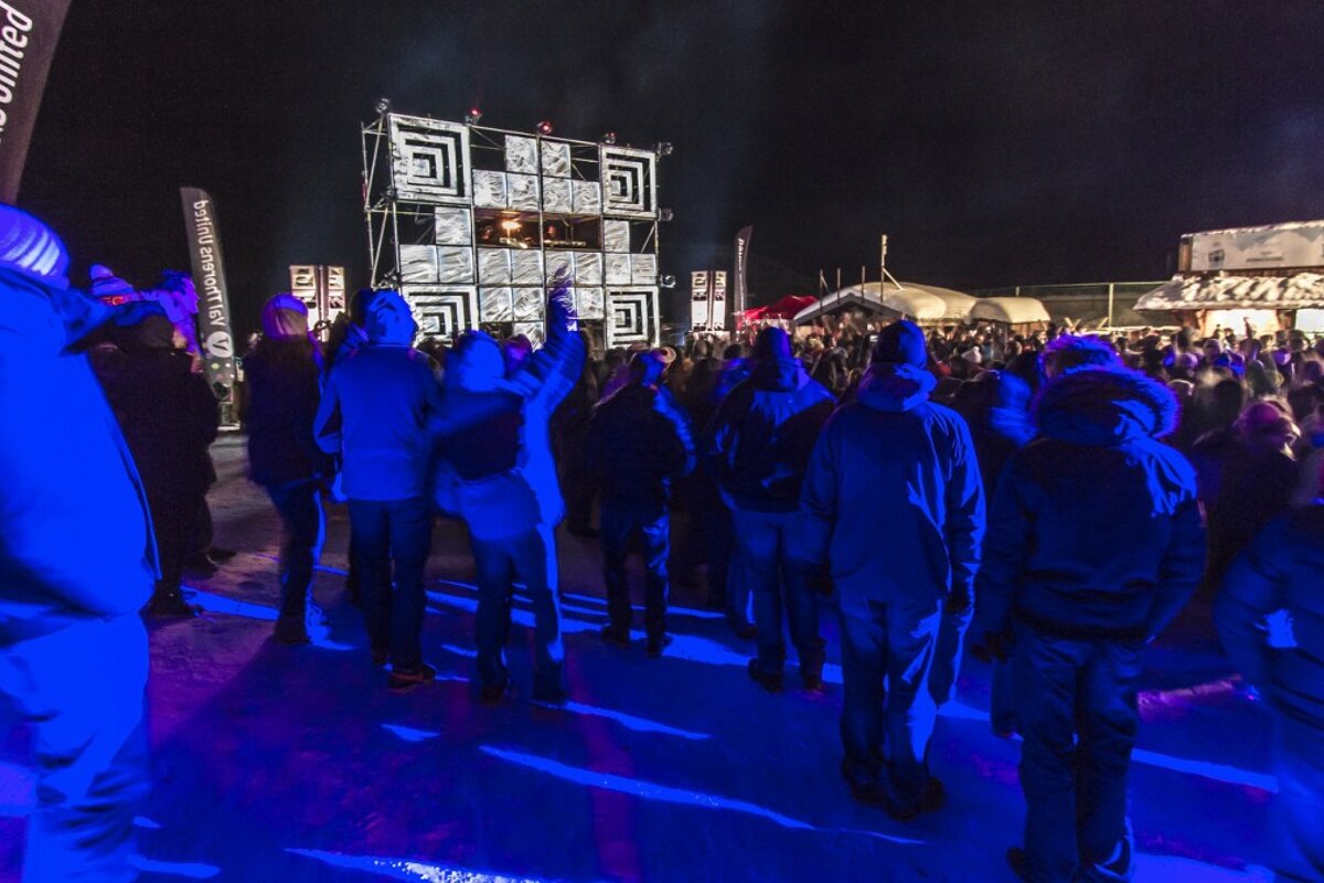 A crowd gathers at an outdoor night concert with a geometric DJ stage and vibrant blue lighting reflecting on the ground.
