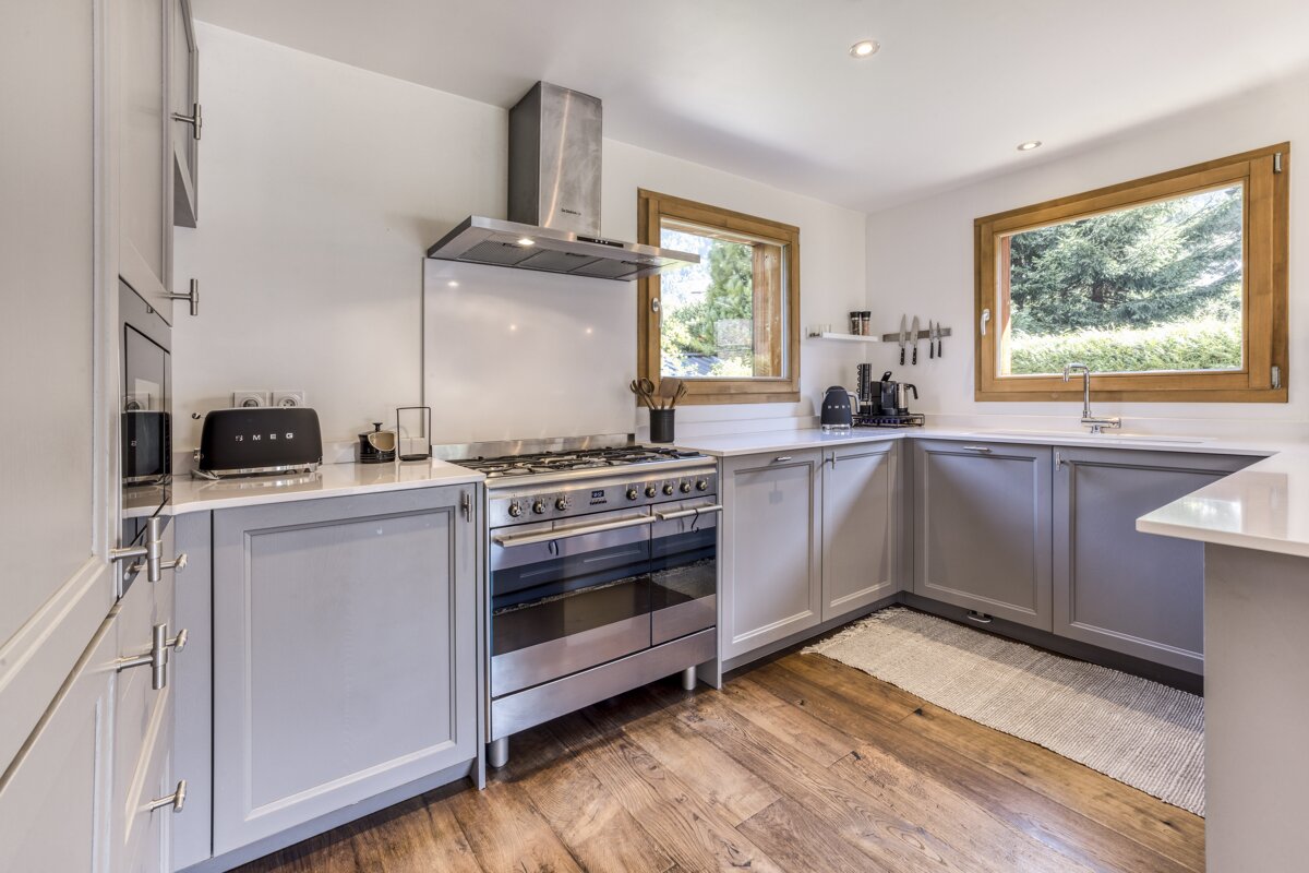 A bright, modern kitchen with light gray cabinets, white countertops, and warm wood floors. Features stainless steel appliances, including a large range, and windows offering greenery views.