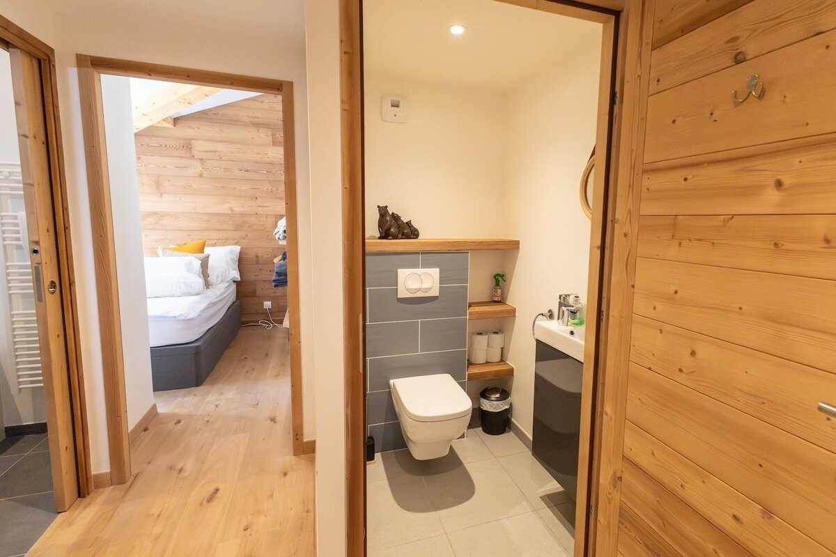 A modern hallway connects a compact bathroom (toilet, sink, wood shelves) on the right with a bedroom (bed, wood paneling) on the left.