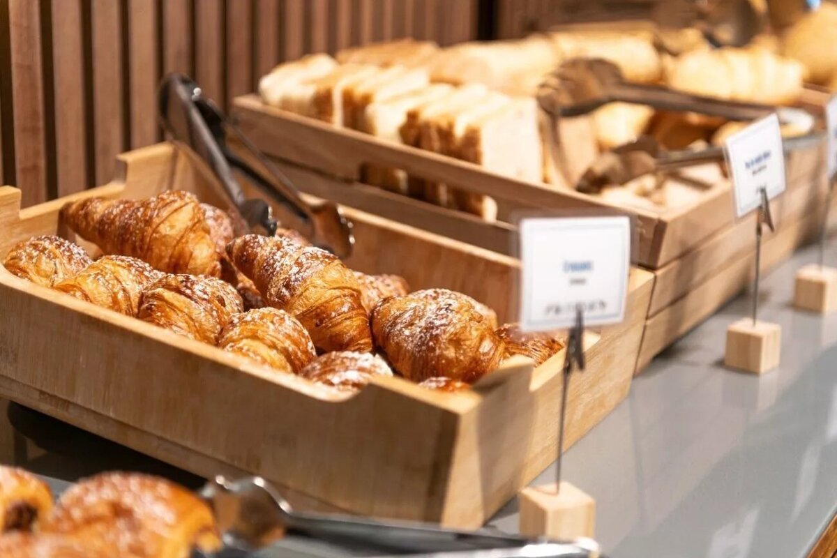 Golden, sugar-dusted croissants fill a wooden buffet tray, with sliced bread behind. Tongs and labels complete this tempting breakfast spread.