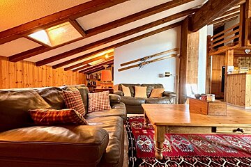 A warm, rustic living room with leather sofas, wooden beams, a red patterned rug, and antique skis decorating a white wall.