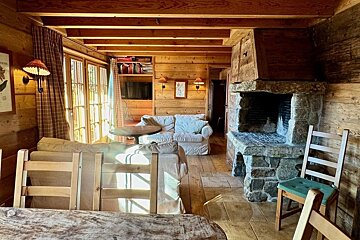 Cozy, wood-paneled cabin living room with a large stone fireplace, exposed ceiling beams, comfortable sofas, and sun streaming through windows.