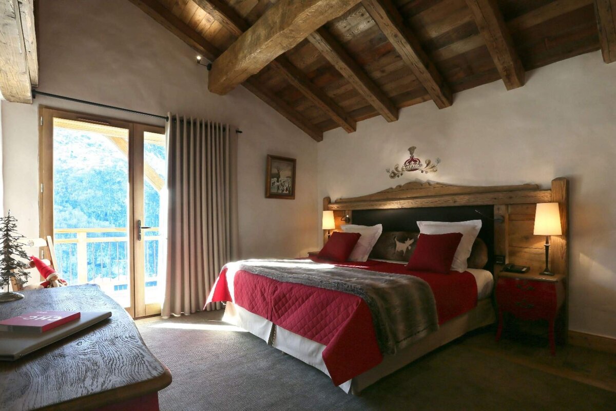 A cozy, rustic bedroom features a large bed with red bedding, a wooden beamed ceiling, and a balcony door revealing a mountain view.
