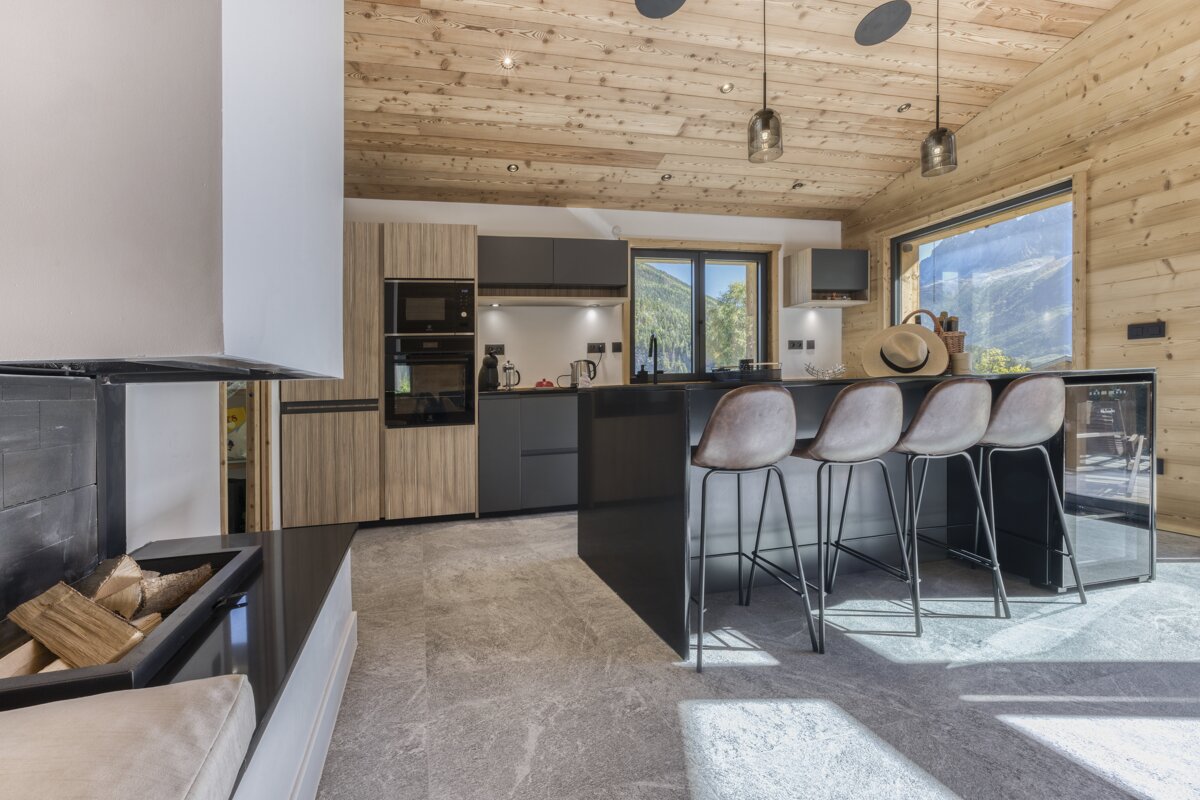 A modern chalet kitchen features light wood paneled walls/ceiling, a black island with bar stools, and a fireplace, all overlooking a stunning mountain view.