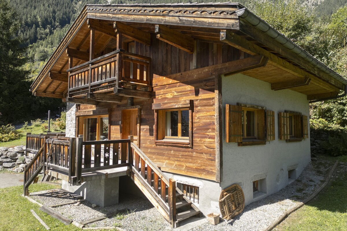 A rustic two-story wooden chalet with a balcony, stairs, and open shutters, set against a lush green, tree-covered hillside on a sunny day.