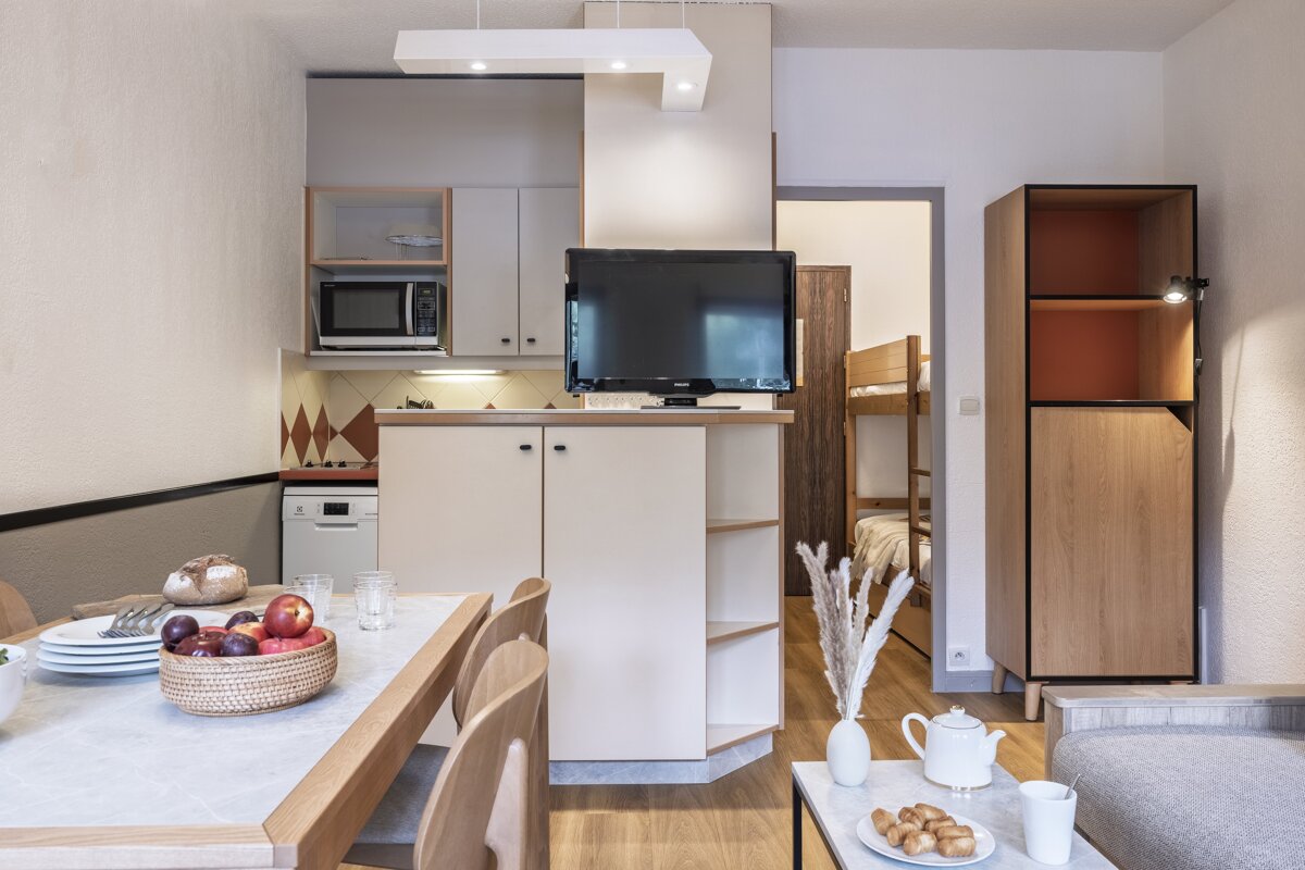 Cozy studio apartment showing a dining area, a kitchen partition with a TV, and a bedroom with bunk beds in the background.