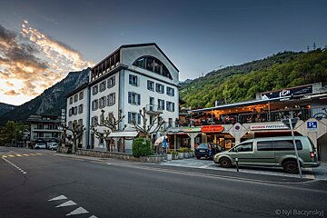 Sunset over a mountain village street with a white hotel, commercial building with a lively restaurant patio, and parked cars.
