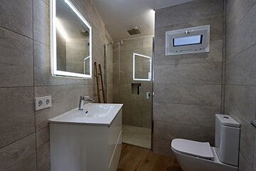 A modern bathroom with grey tiled walls, a white vanity and illuminated mirror, a glass shower, toilet, and wooden floor.