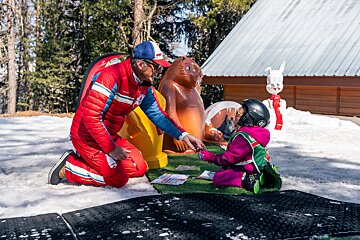A ski instructor kneels in the snow, interacting with a young child in a pink ski suit and helmet, with cartoon animal figures nearby.