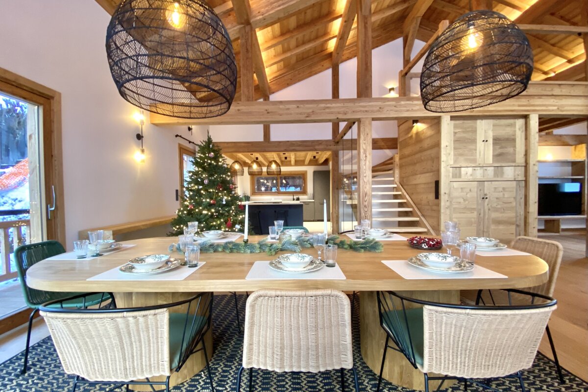 A cozy, rustic dining room with a set table, timber beams, and a Christmas tree. Woven pendant lights illuminate the festive space.