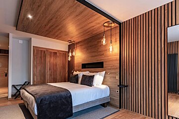 A modern bedroom with warm wood paneling on walls and ceiling, a bed with white and dark bedding, and stylish Edison bulb pendant lights.