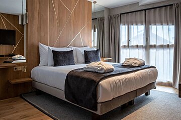 Modern hotel room with a large bed, wooden headboard with gold accents, two bathrobes, and a window with sheer curtains.